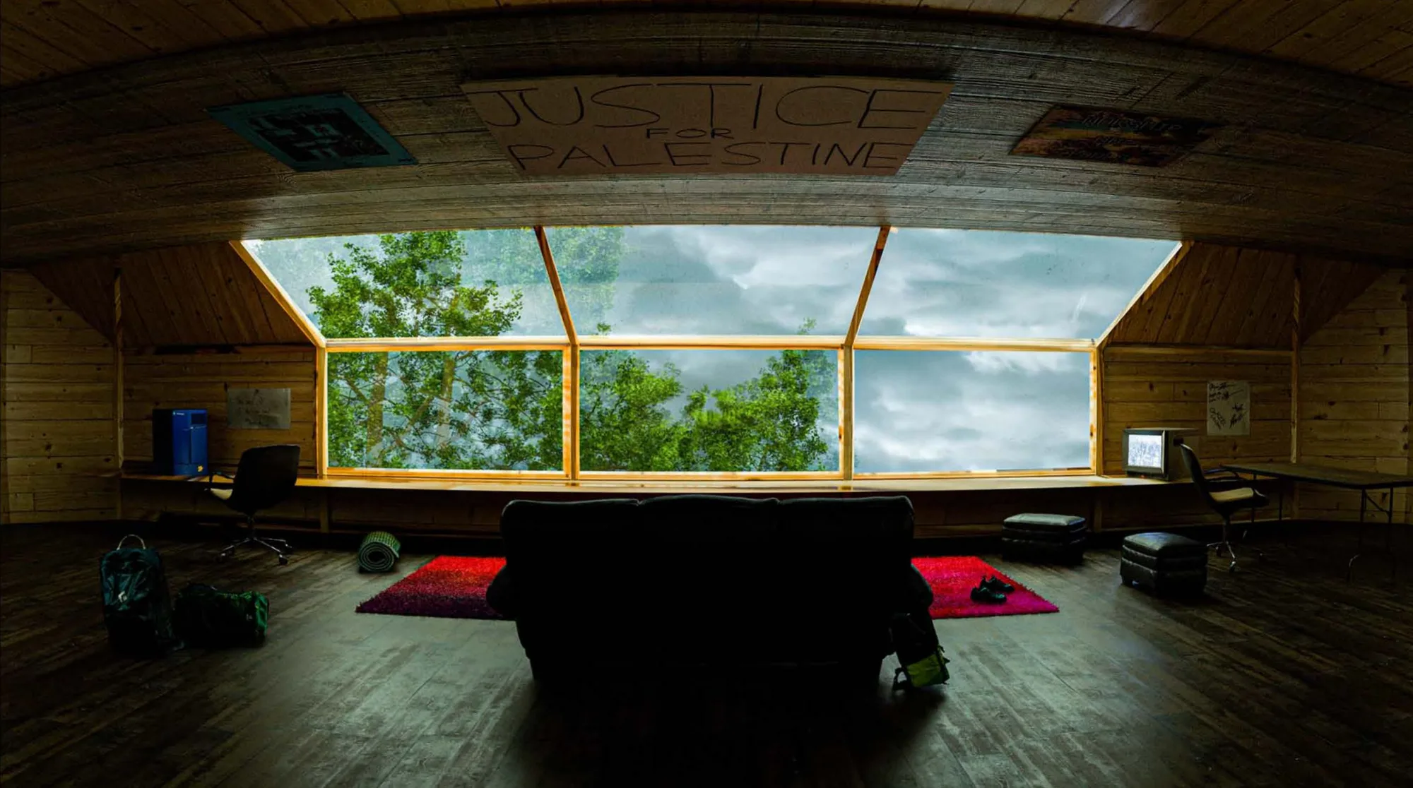 A wooden cabin interior with a wide window looking out on trees.